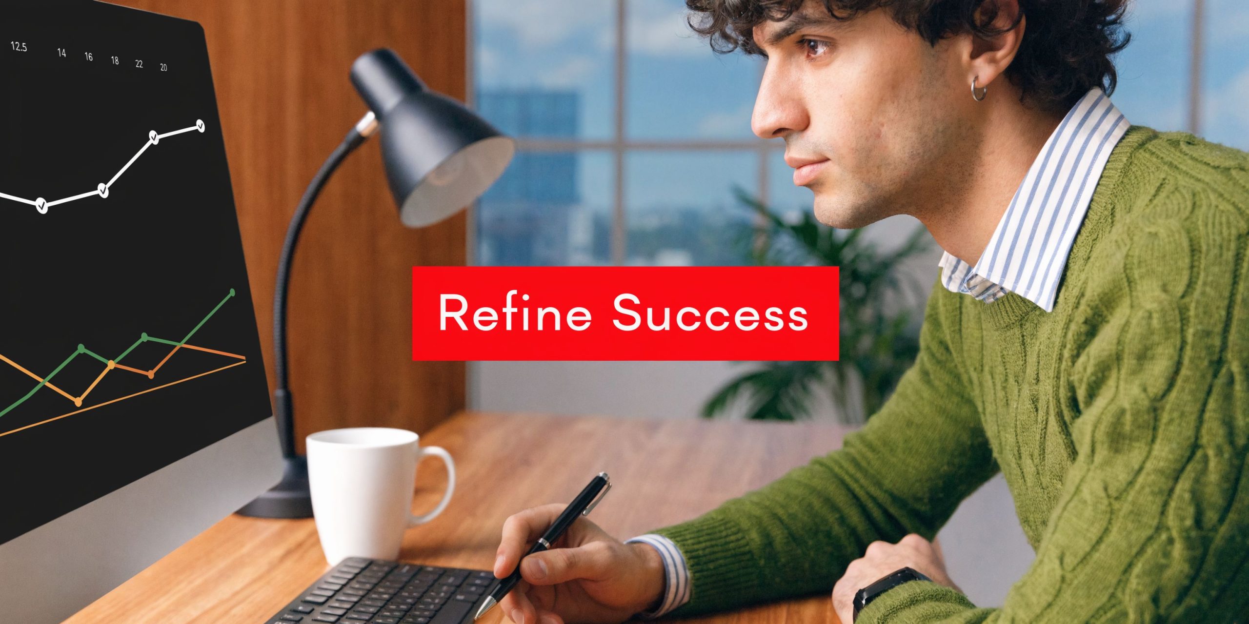 A young man sitting at a wooden desk with a computer, writing notes while observing a business chart.