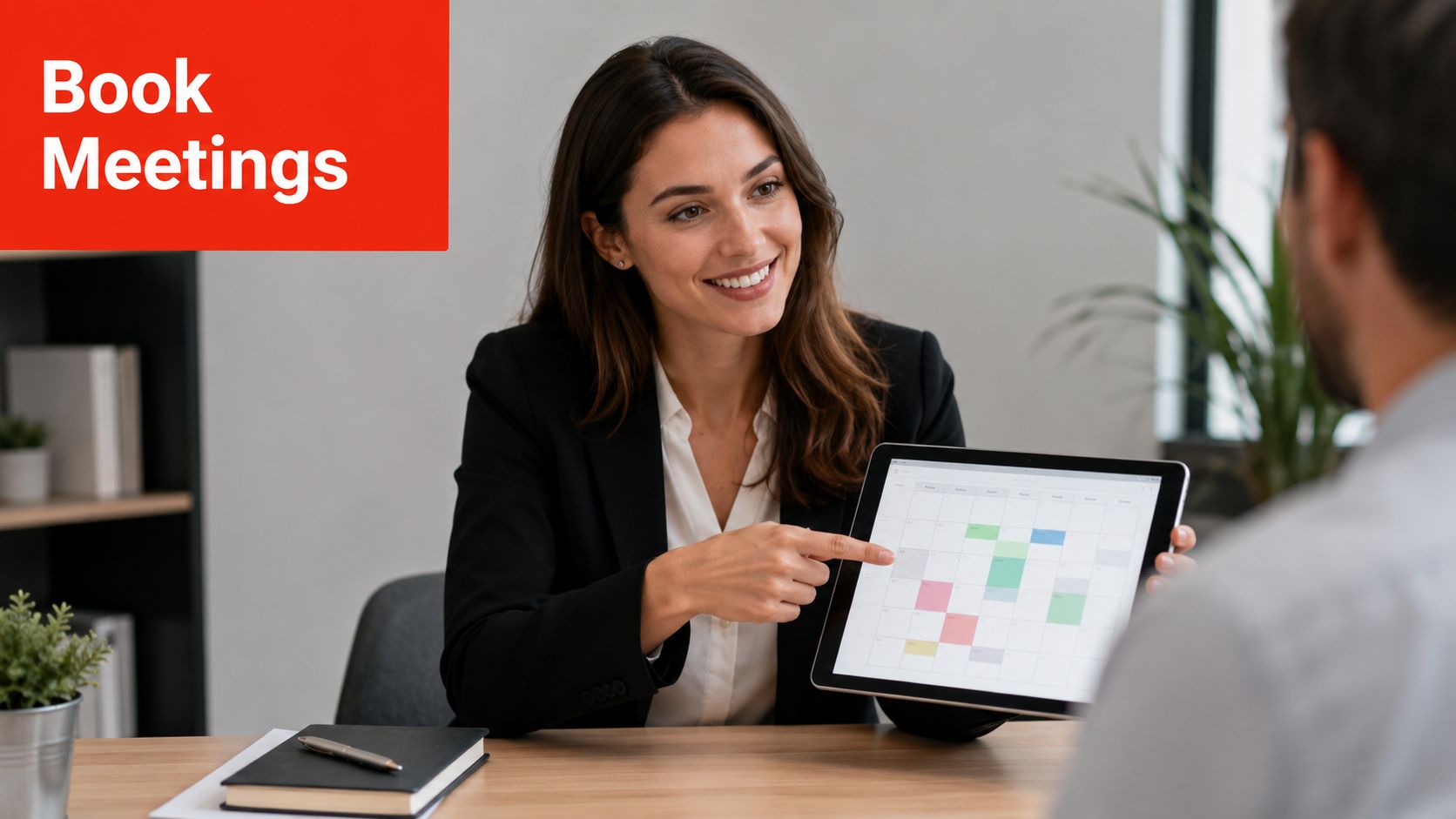 A smiling businesswoman in a professional suit shows a digital calendar on a tablet to a colleague.